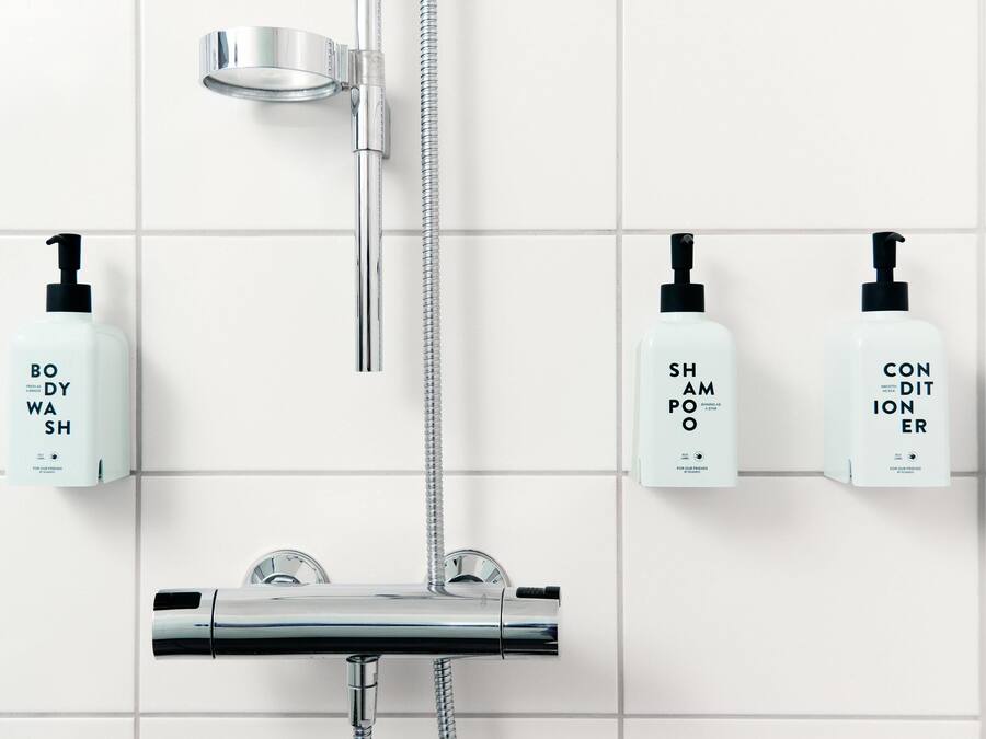 A bathroom with a chrome showerhead, a handheld showerhead, and three bottles of body wash, shampoo, and conditioner on the wall.