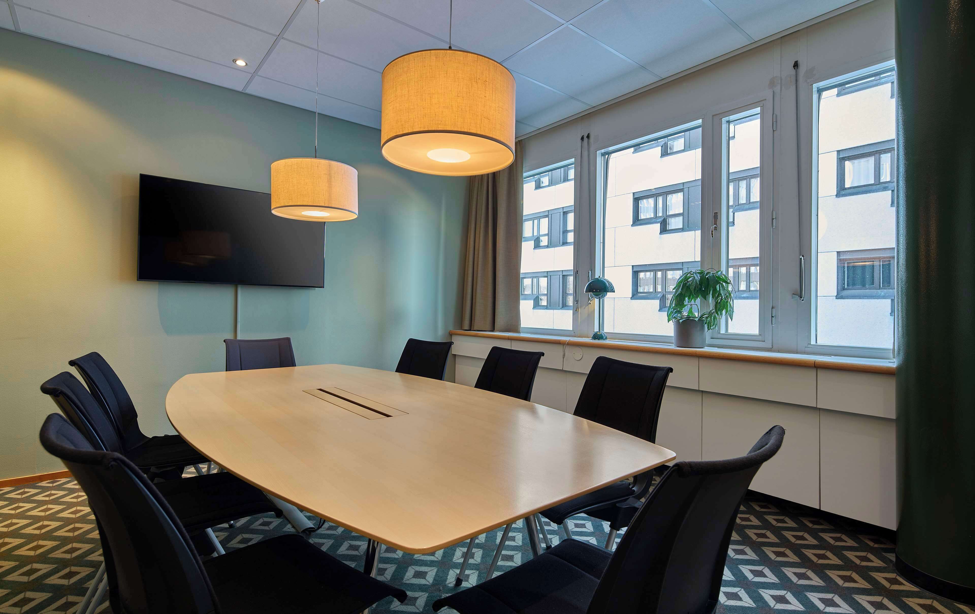 A conference room with a large table, chairs, a TV, and a window with a view of buildings.