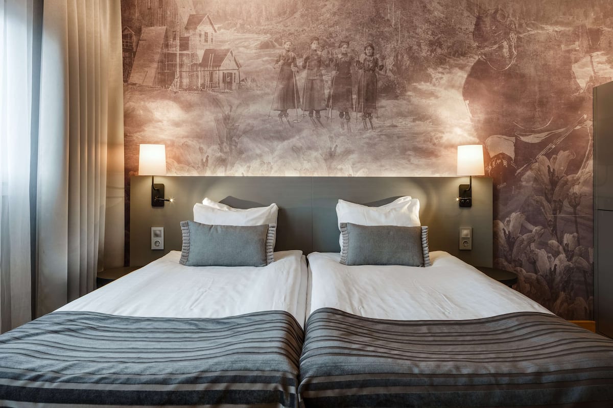 Uma cama de casal com duas almofadas, cabeceira com painel decorativo e duas luminárias fixadas na parede.