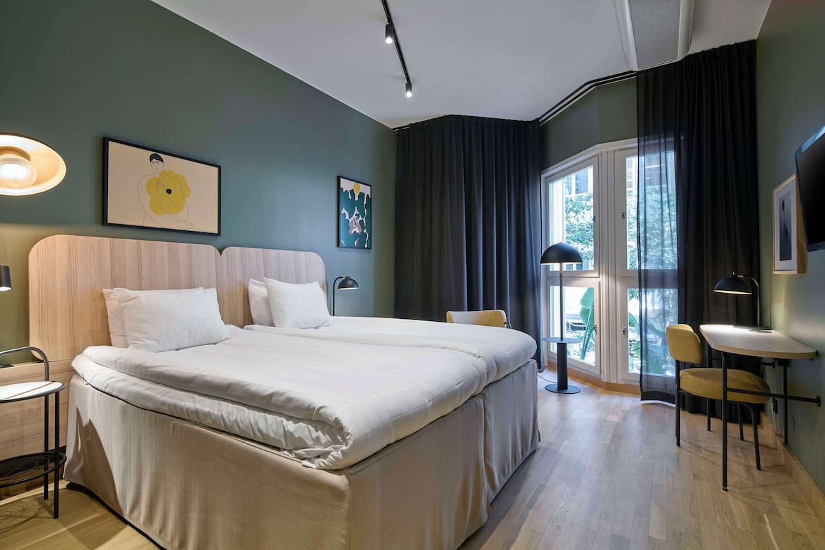 Quarto de hotel moderno com uma cama grande, uma mesa pequena, uma cadeira e uma janela com cortinas.