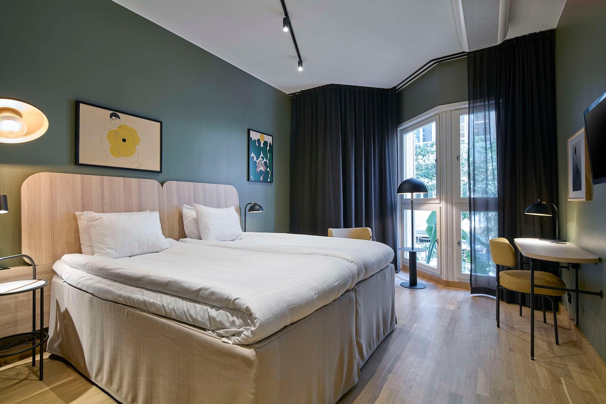 Habitación de hotel moderna con una cama grande, un escritorio y una silla. Hay dos cuadros en la pared y una ventana con cortinas.