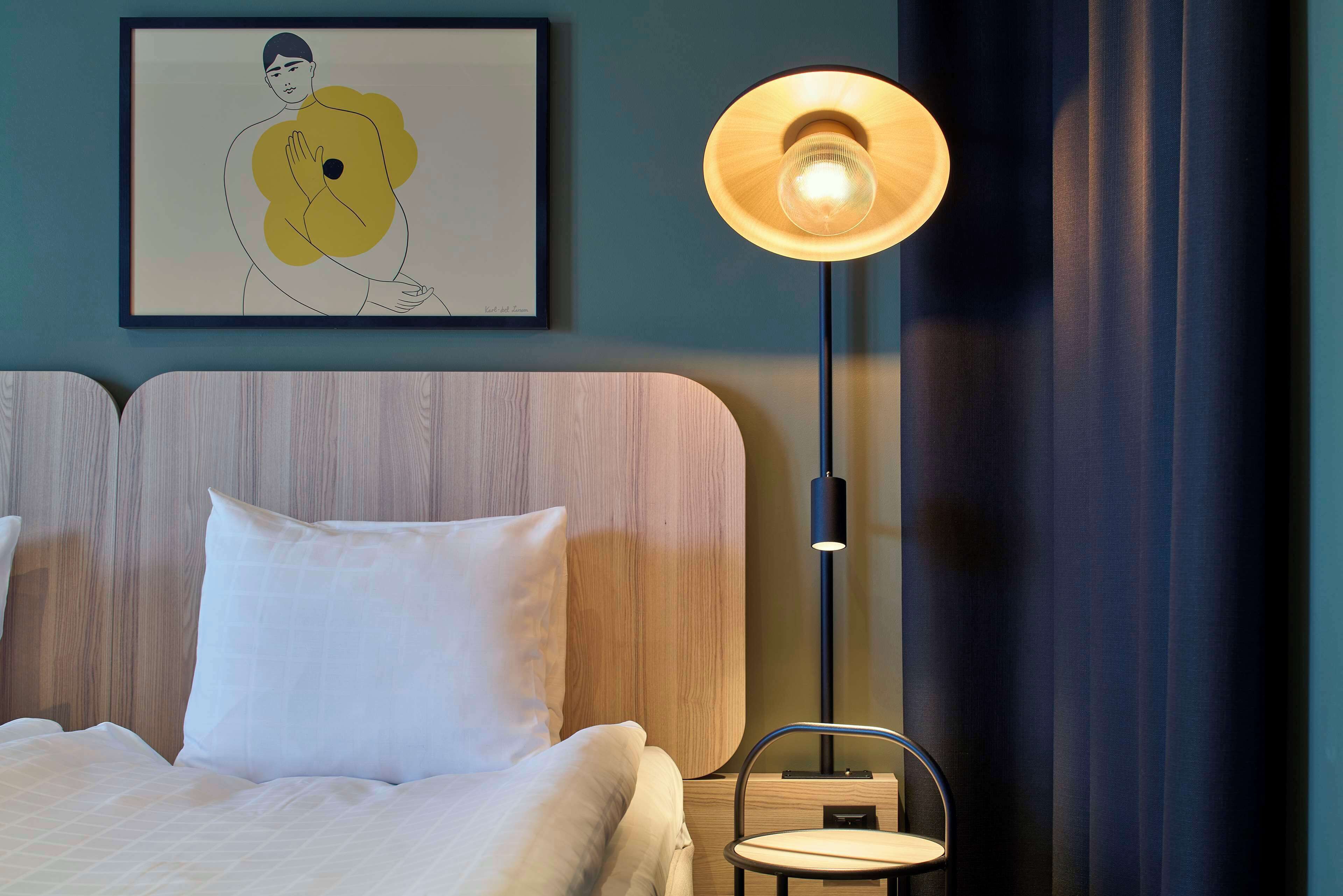 Una cama bien hecha con ropa blanca, una obra de arte enmarcada en la pared y una lámpara de pie con pantalla de madera.