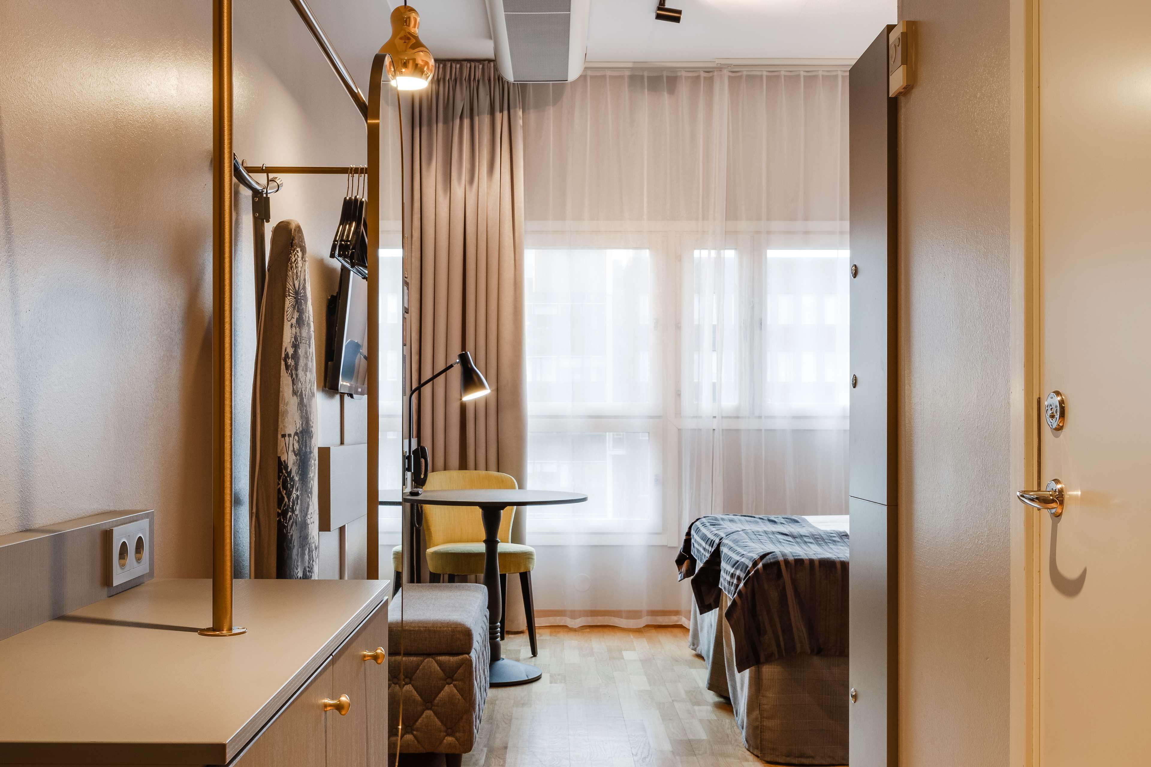 A compact hotel room with a bed, a small table, a chair, a desk, and a wardrobe.
