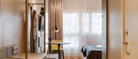 A compact hotel room with a bed, a small table, a chair, a desk, and a wardrobe.