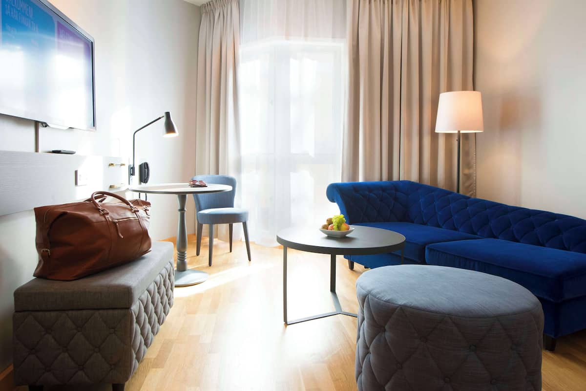 Quarto de hotel moderno com sofá azul, um banquinho cinza, uma pequena mesa redonda com abajur e uma grande janela com cortinas.