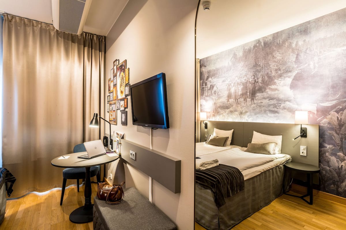 Quarto de hotel com uma cama, uma escrivaninha com um laptop, uma televisão e um quadro na parede.