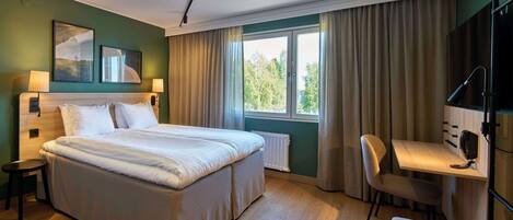 A hotel room with a large bed, a desk with a chair, a window with curtains, and a radiator.