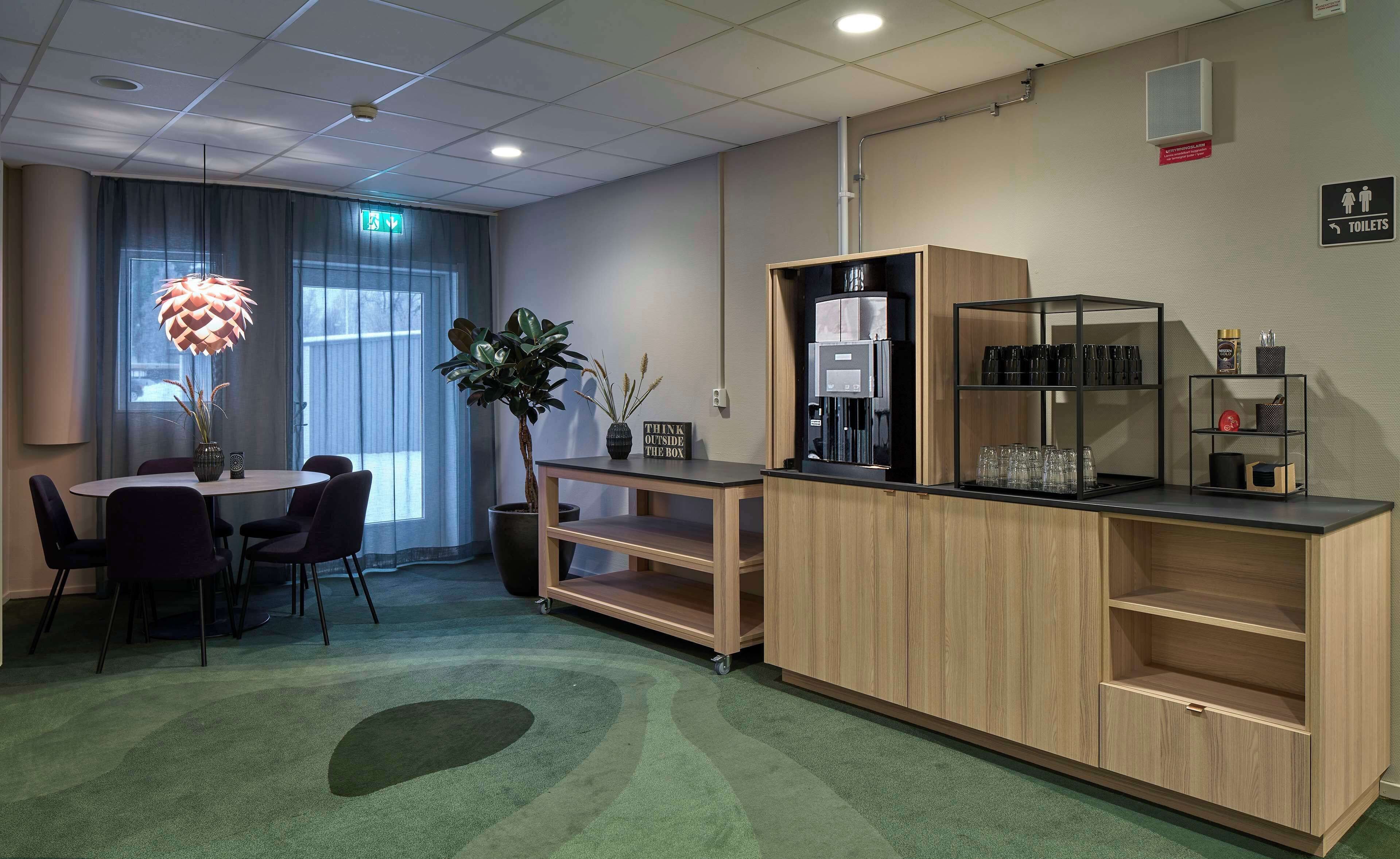 Una habitación de hotel moderna con zona de comedor, cafetera y estante con vasos.