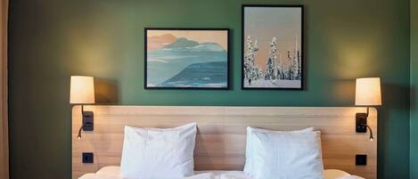 Cama doble con ropa blanca, dos lámparas fijadas a la pared y dos cuadros de paisajes en una pared verde.