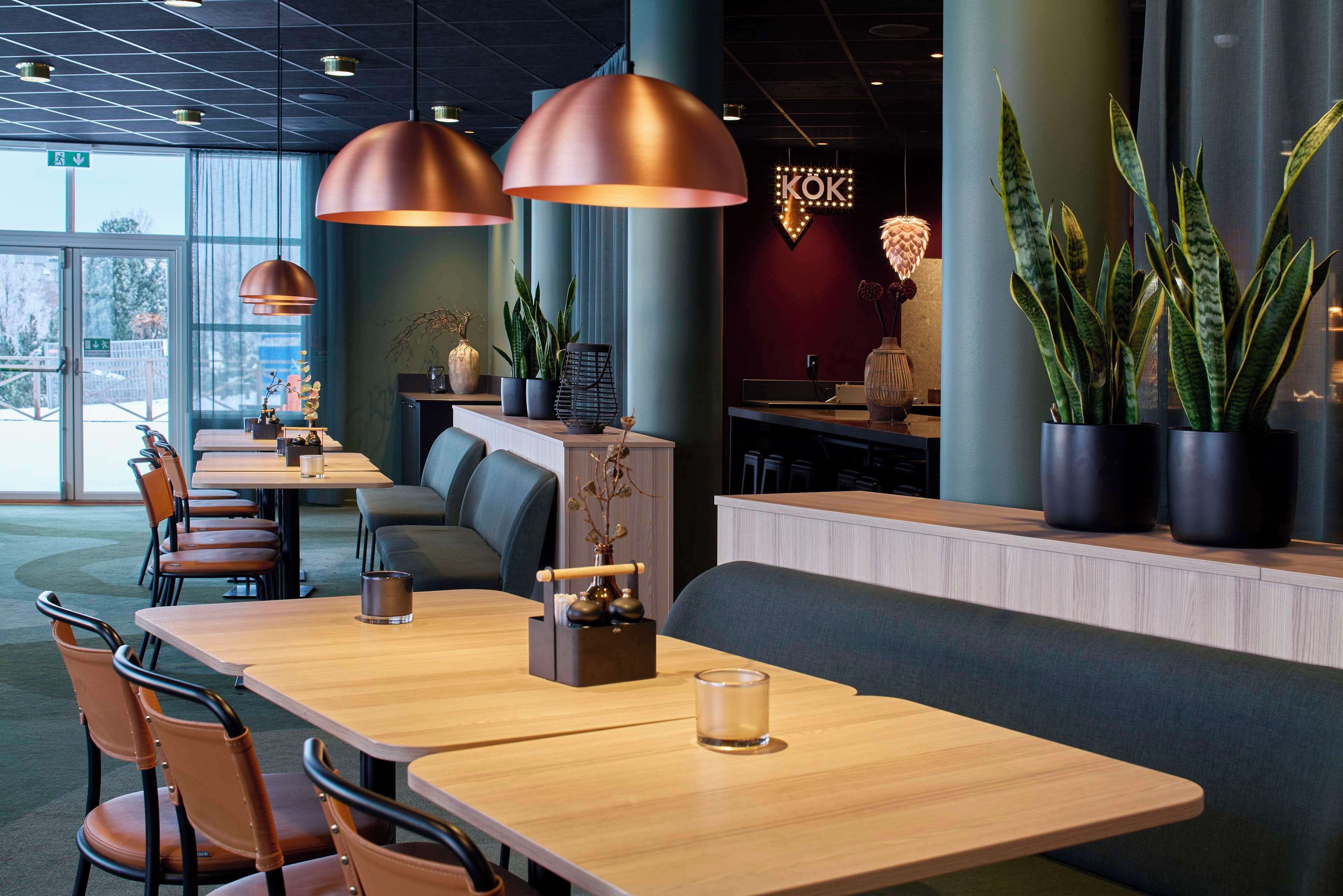 Un restaurant moderne avec des tables et des chaises en bois, des luminaires suspendus, des plantes en pot et un panneau lumineux au néon.