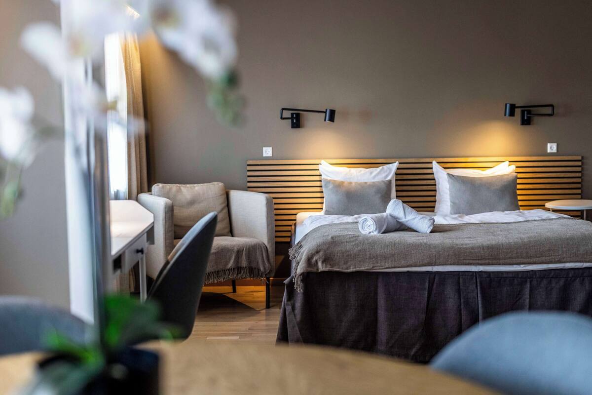 A modern hotel room with a bed, a desk, and a chair. There are wall-mounted lights above the bed and a small round table beside it.