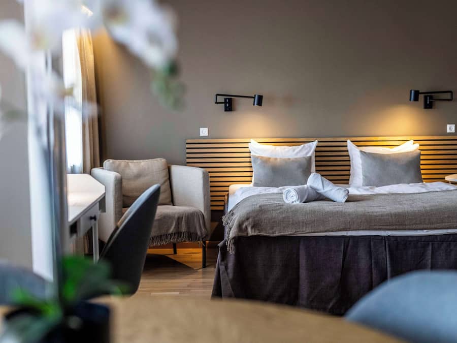 A modern hotel room with a bed, a desk, and a chair. There are wall-mounted lights above the bed and a small round table beside it.