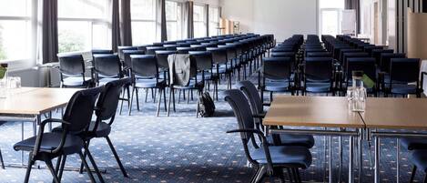 Une salle de conférence avec des rangées de chaises et de tables, de grandes fenêtres avec des rideaux et un écran de projection.