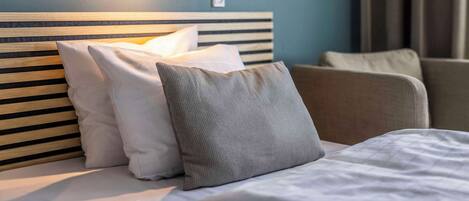 Habitación de hotel con cabecera de madera, almohadas blancas y grises, una lámpara de pared y vista a unas cortinas.
