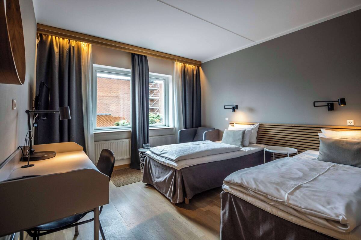 A hotel room with two beds, a desk, a chair, a small table, and a window with curtains.