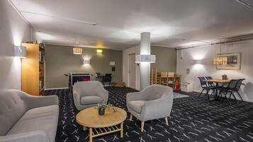 Una habitación de hotel moderna con alfombra de estampado blanco y negro, dos sillones grises, una mesa redonda de madera, una estantería y un comedor con mesa y sillas.