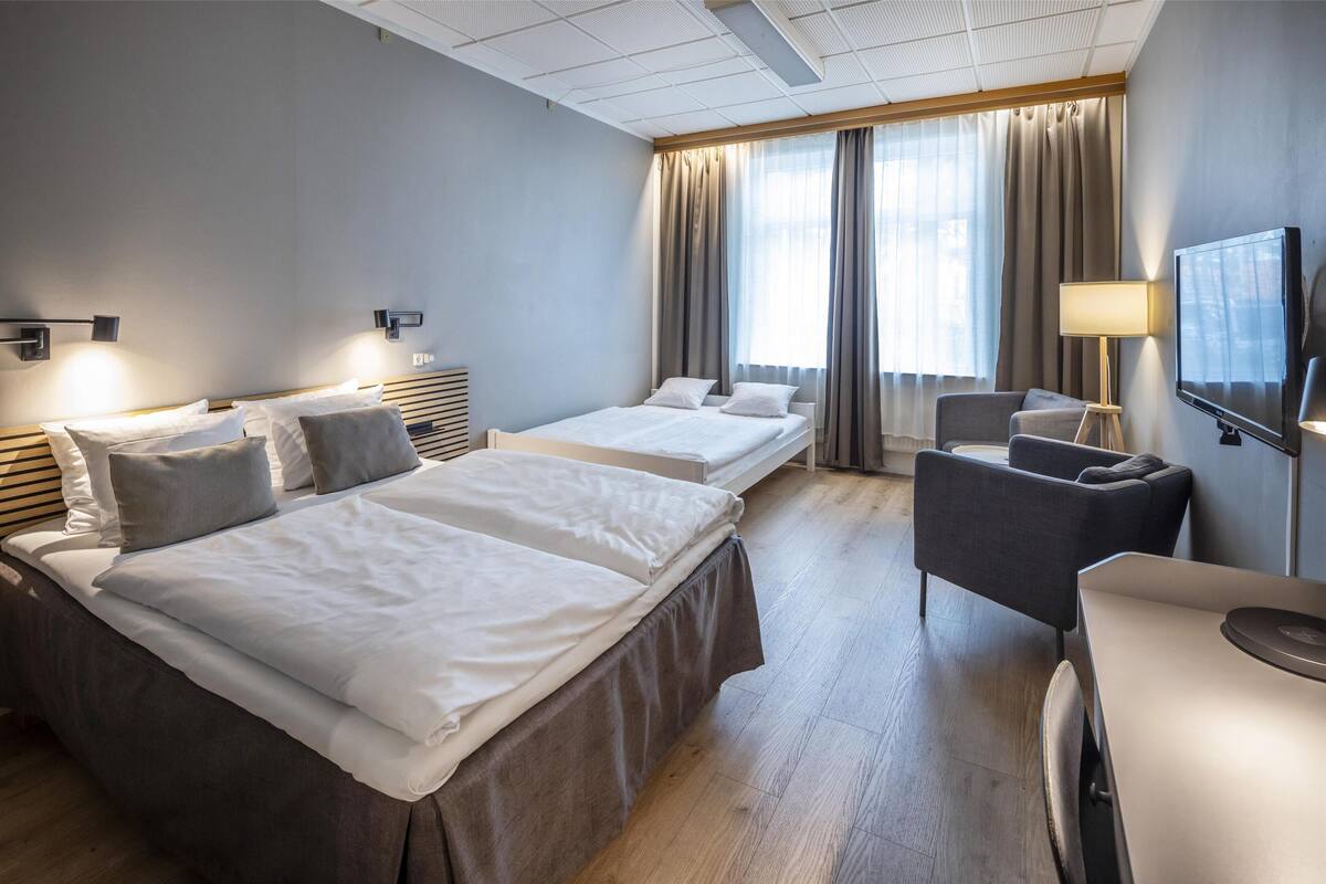 A hotel room with two beds, a desk, a chair, a TV, and a large window with curtains.
