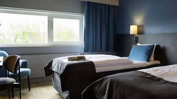 A hotel room with two beds, a window with curtains, a chair, and a lamp.