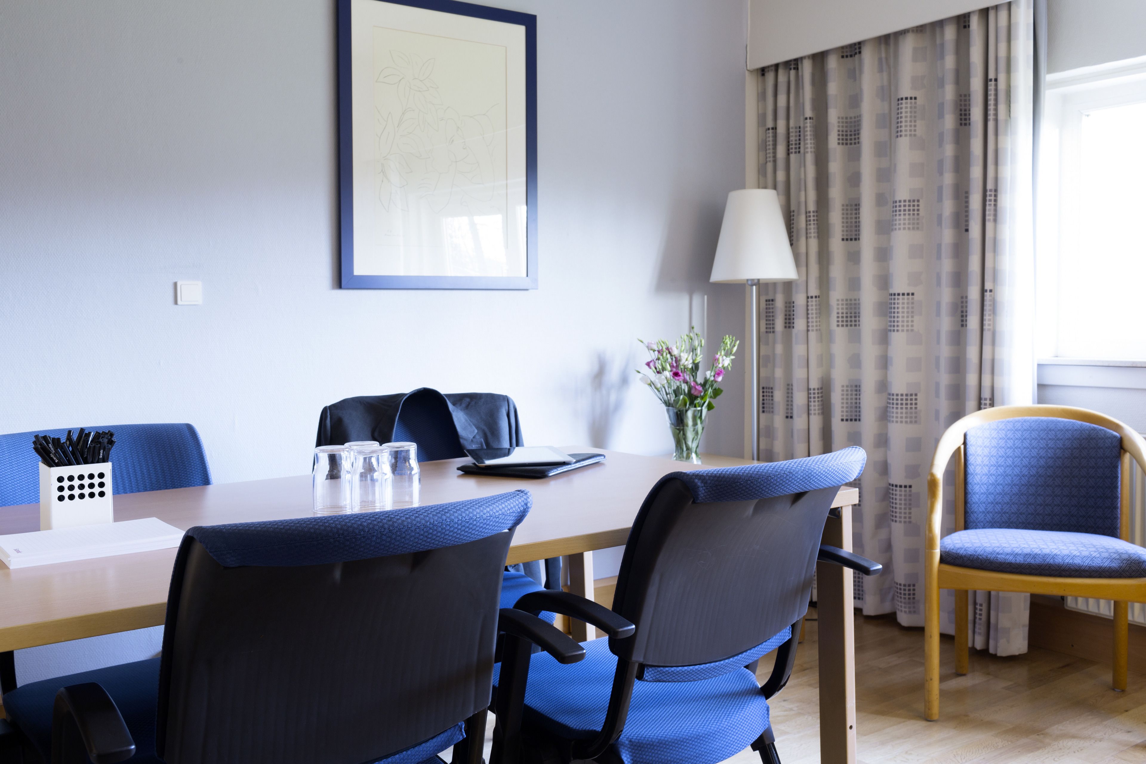 Une salle de conférence avec une table en bois, des chaises bleues, une lampe blanche et une œuvre d’art encadrée au mur.