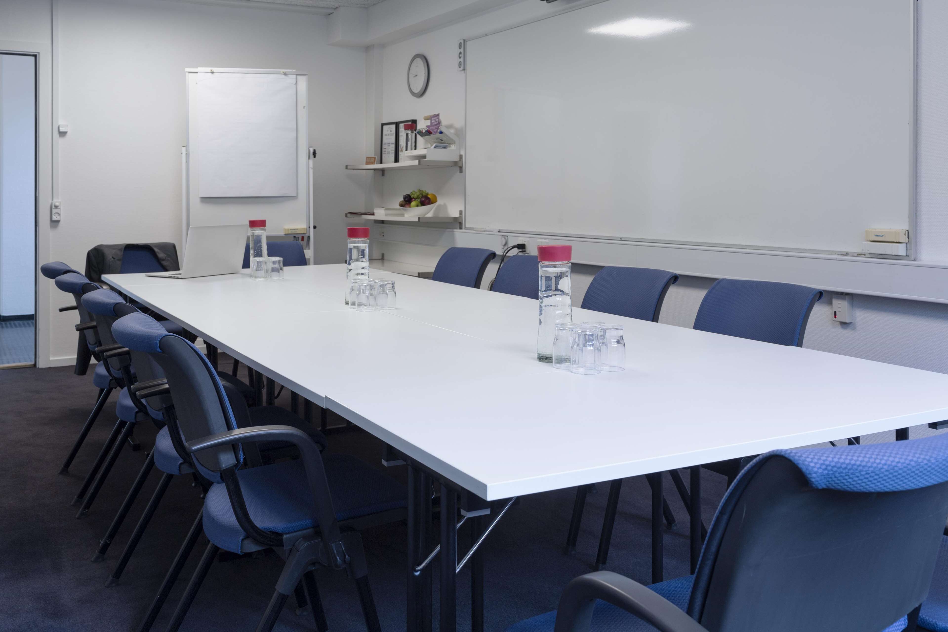 Un salón de conferencias con una larga mesa blanca, sillas azules, botellas de agua, una pizarra y estantes con libros y fruta.