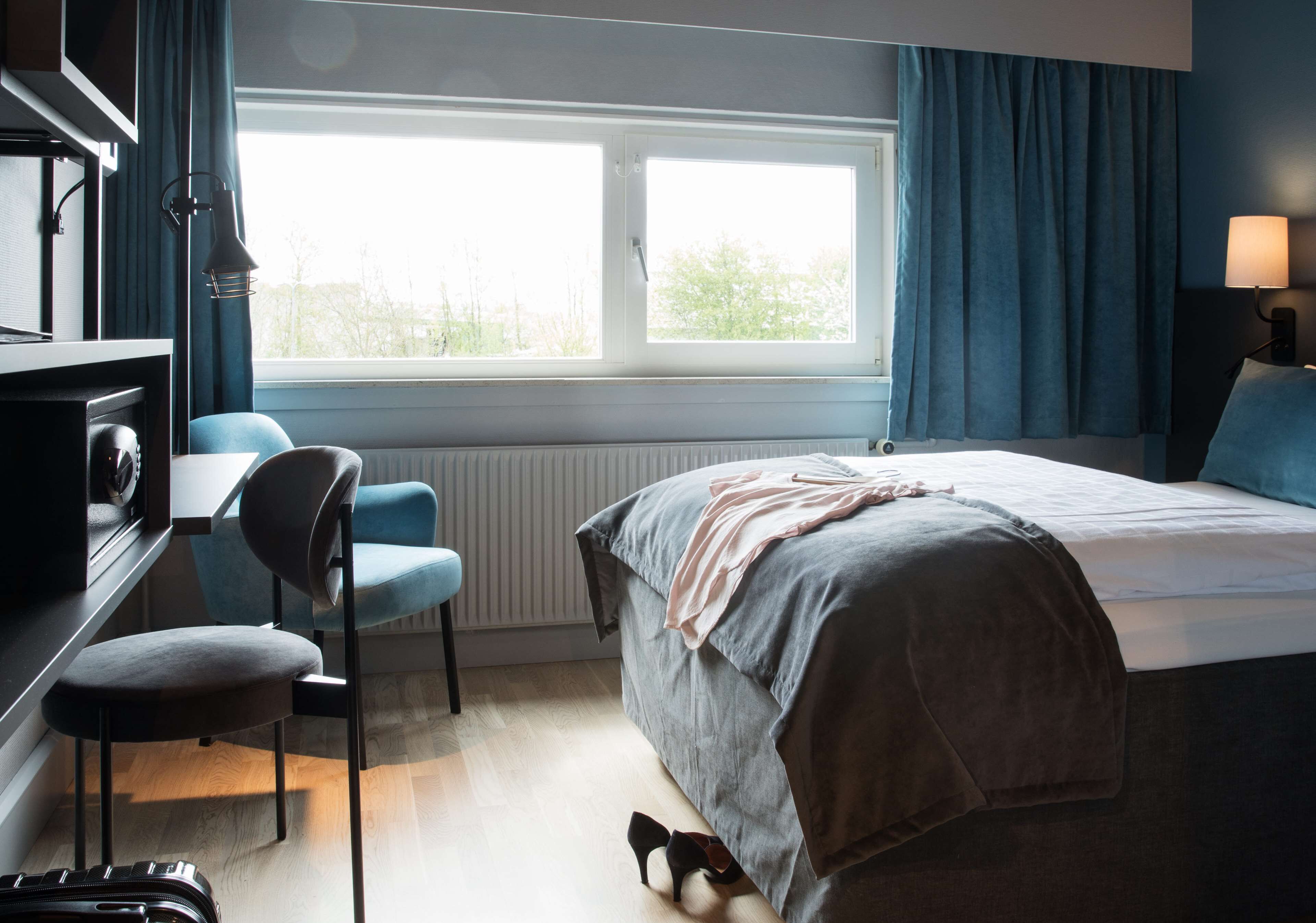 Et hotelværelse med en seng, et skrivebord, en stol, et vindue og en lampe.