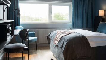 A hotel room with a bed, a desk, a chair, a window, and a lamp.