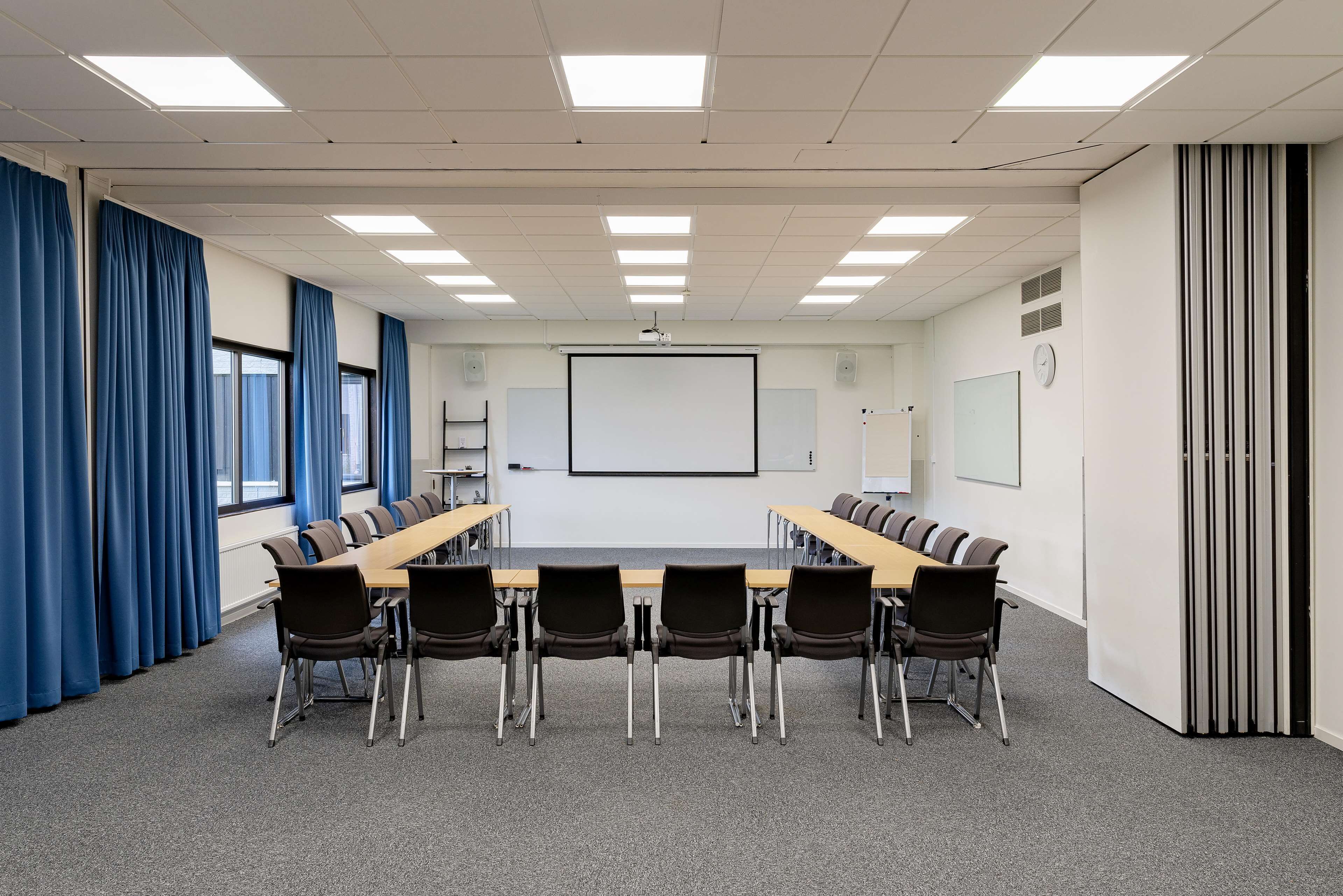 A conference room with a long table, six chairs, a projector screen, a whiteboard, and a clock on the wall.