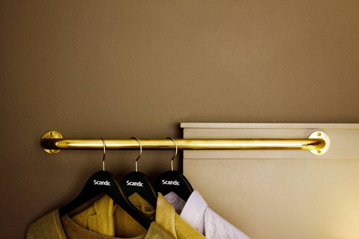 A brass curtain rod with three black hangers labeled "Scandic" against a beige wall.