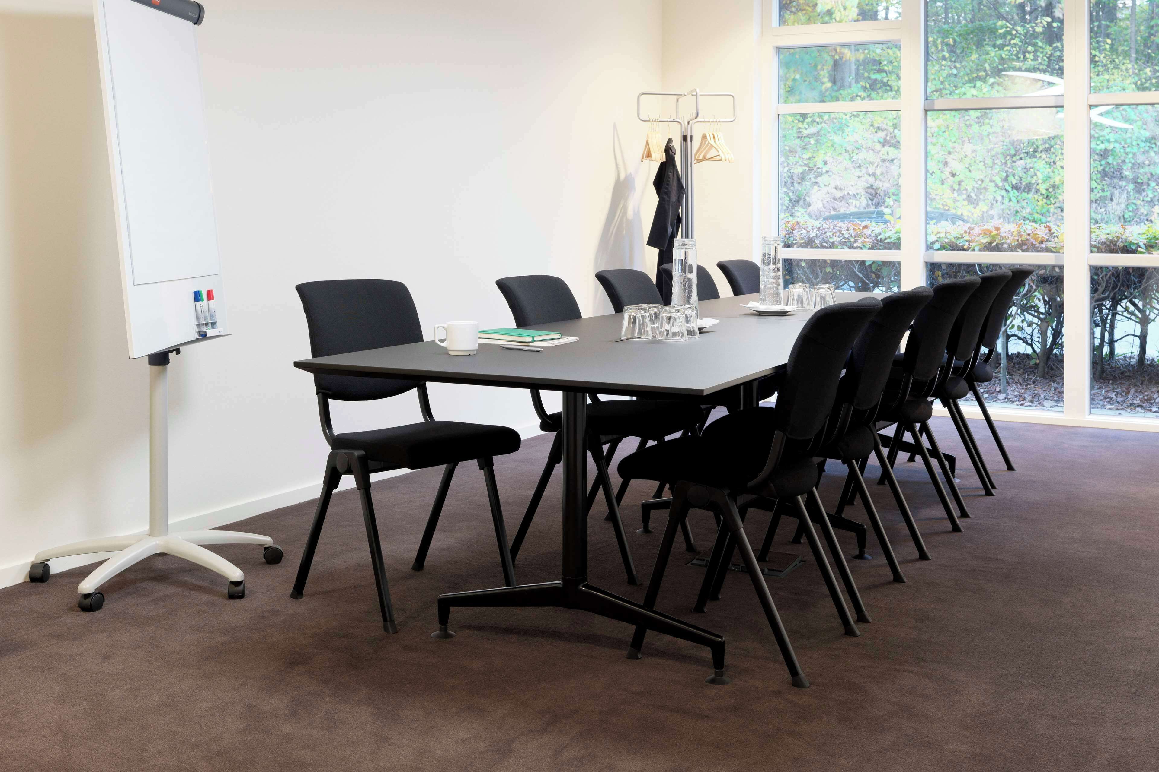A conference room with a large table, black chairs, a whiteboard, and a view of trees through large windows.