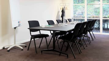 Een vergaderruimte met een grote tafel, zwarte stoelen, een whiteboard en uitzicht op bomen door grote ramen.