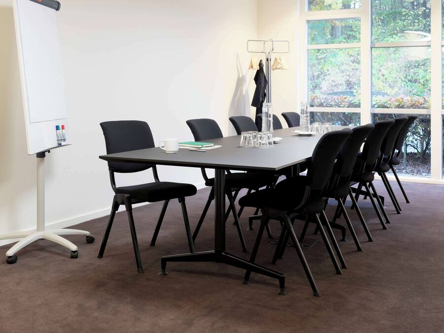 Een vergaderruimte met een grote tafel, zwarte stoelen, een whiteboard en uitzicht op bomen door grote ramen.