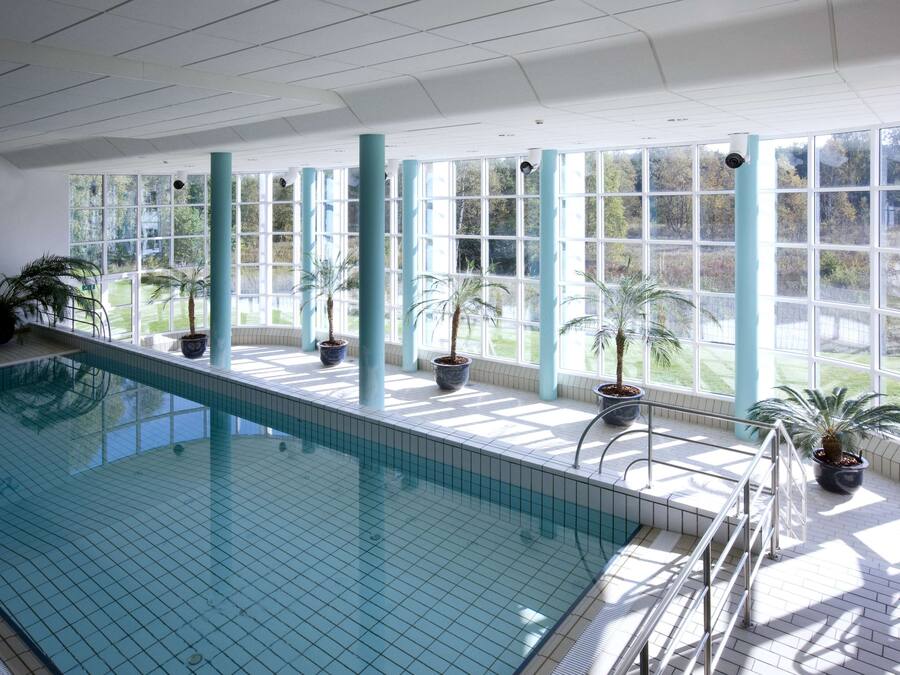 An indoor swimming pool with clear glass walls, potted palm trees, and a view of a green landscape.