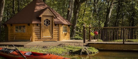 Una cabaña de troncos con puerta y ventanas de madera, una canoa roja con remo, y un pequeño puente sobre un cuerpo de agua tranquilo.