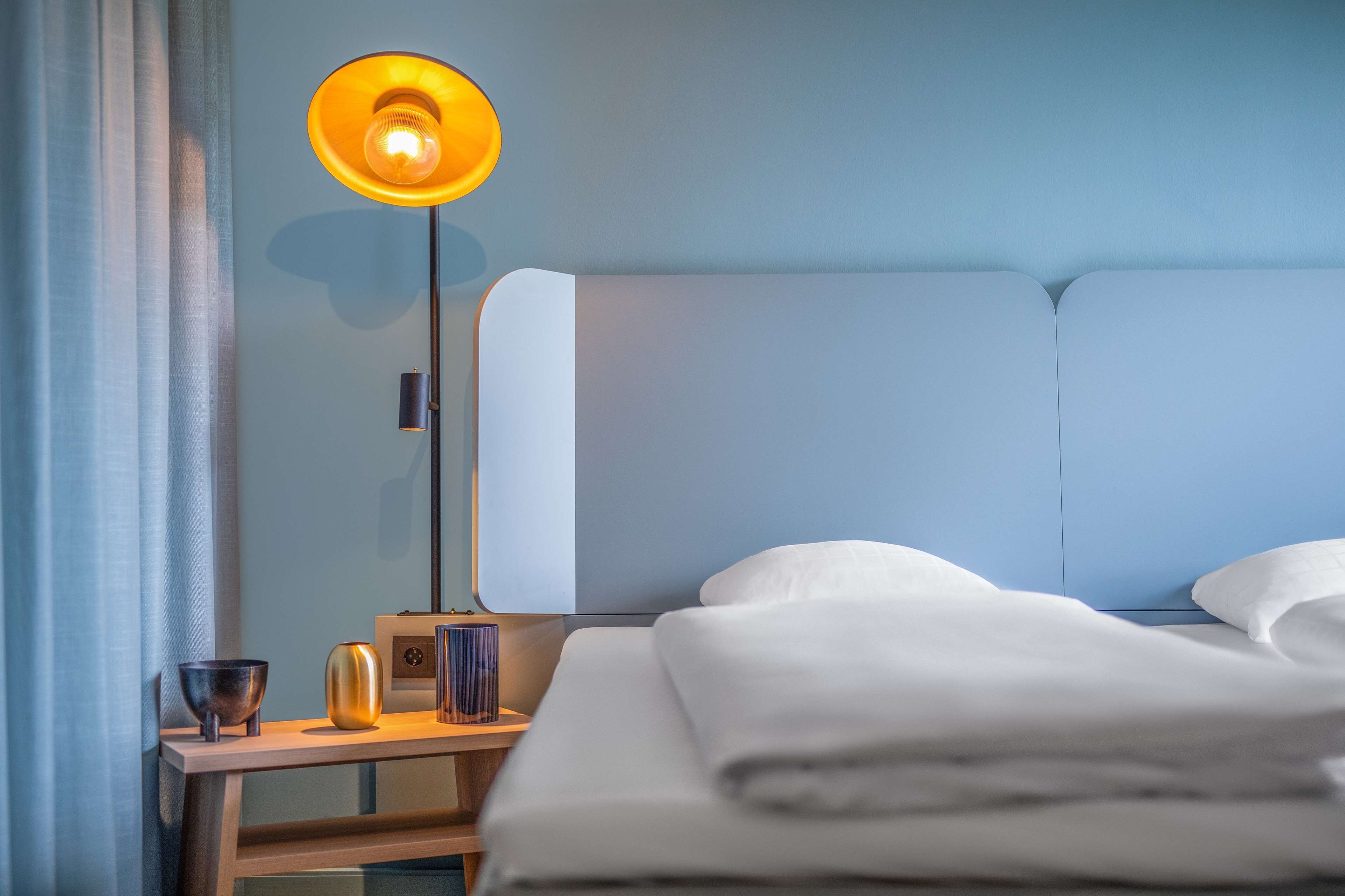 En pænt redt seng med hvide sengetæpper, et natbord med lampe, skål og vase.
