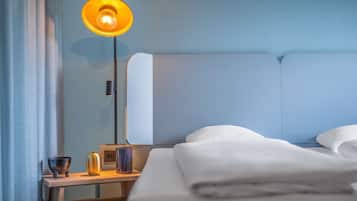 En pænt redt seng med hvide sengetæpper, et natbord med lampe, skål og vase.