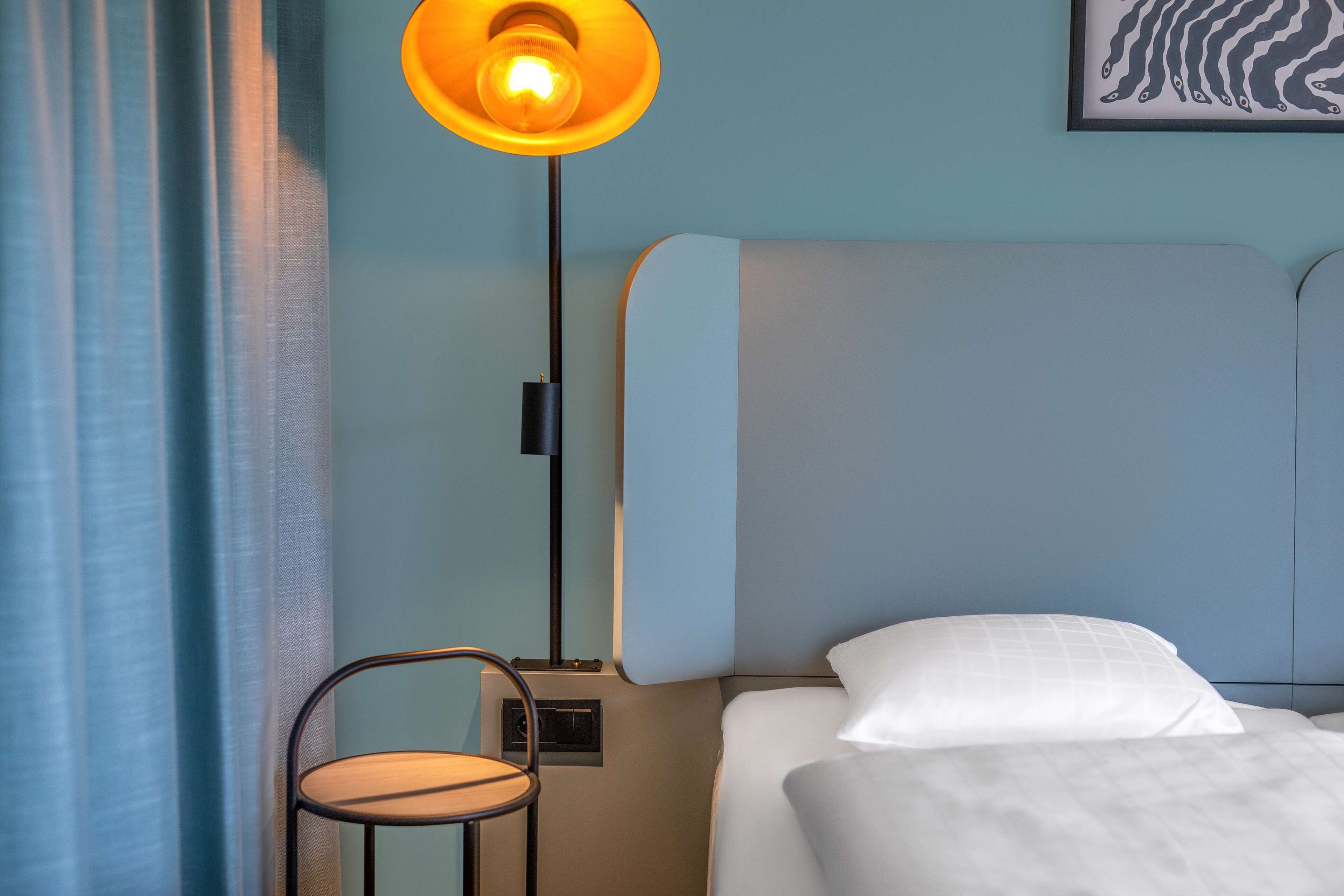 A neatly made bed with white linens, a single chair, a floor lamp, and a framed artwork on the wall.