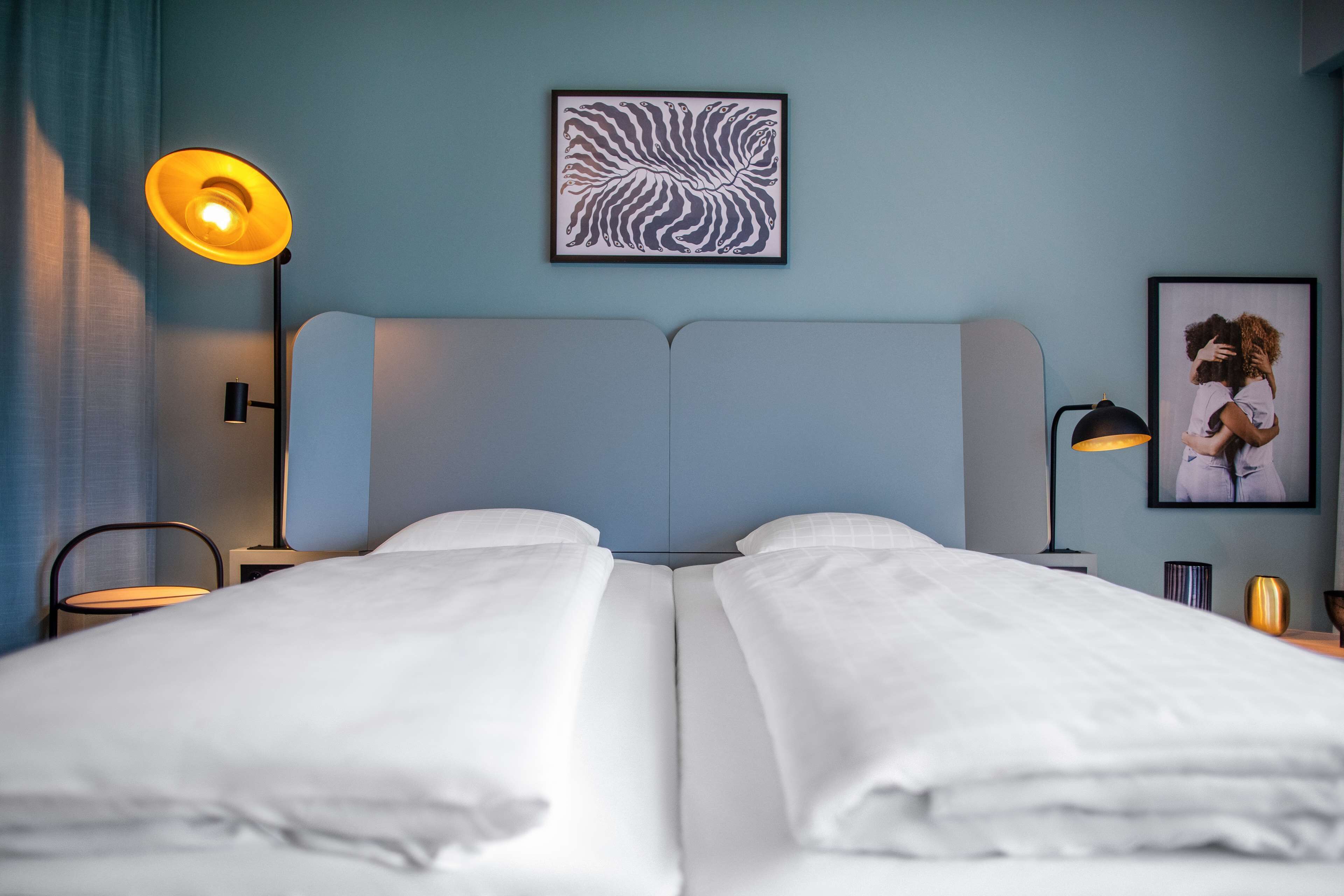 Una cama doble con ropa de cama blanca, un cabecero, una obra de arte abstracto colgada en la pared, una fotografía enmarco y una lámpara de pie.