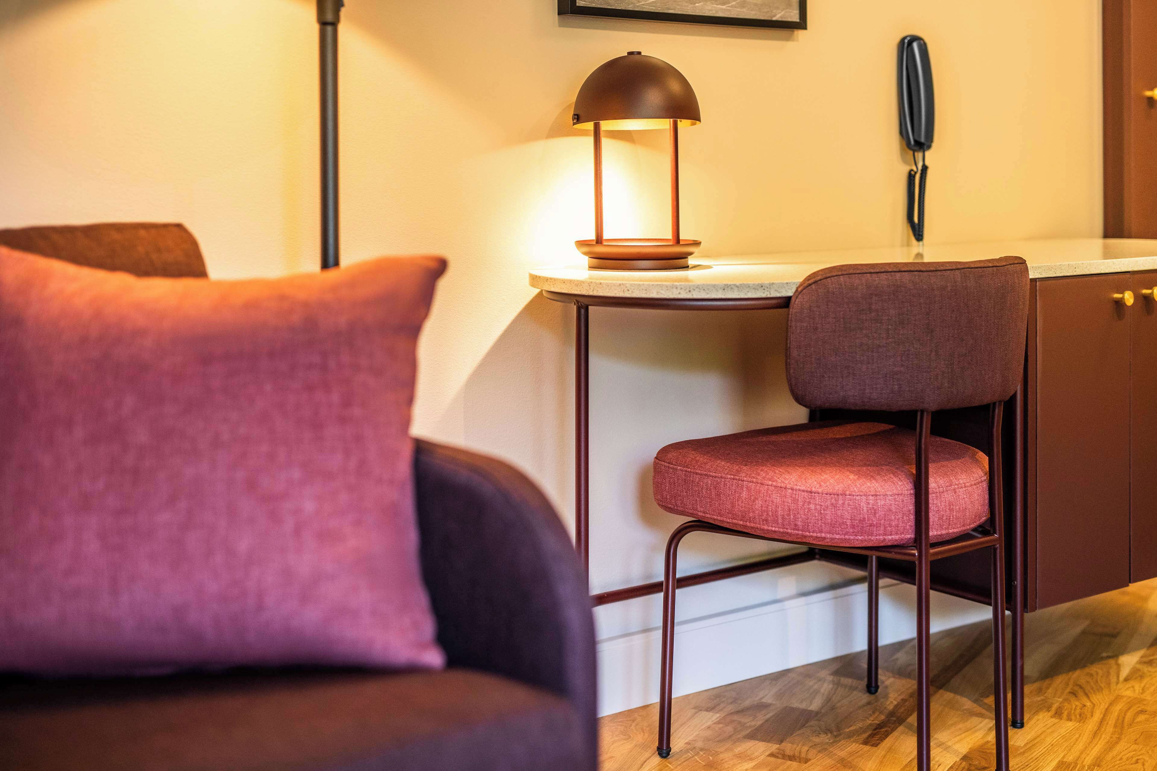 Una habitación moderna con un sofá de color oscuro, un sillón rojo acolchado, una pequeña mesa redonda con una lámpara y un teléfono en la pared.