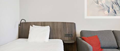 A hotel room with a bed, a gray sofa, and a wall-mounted control panel.