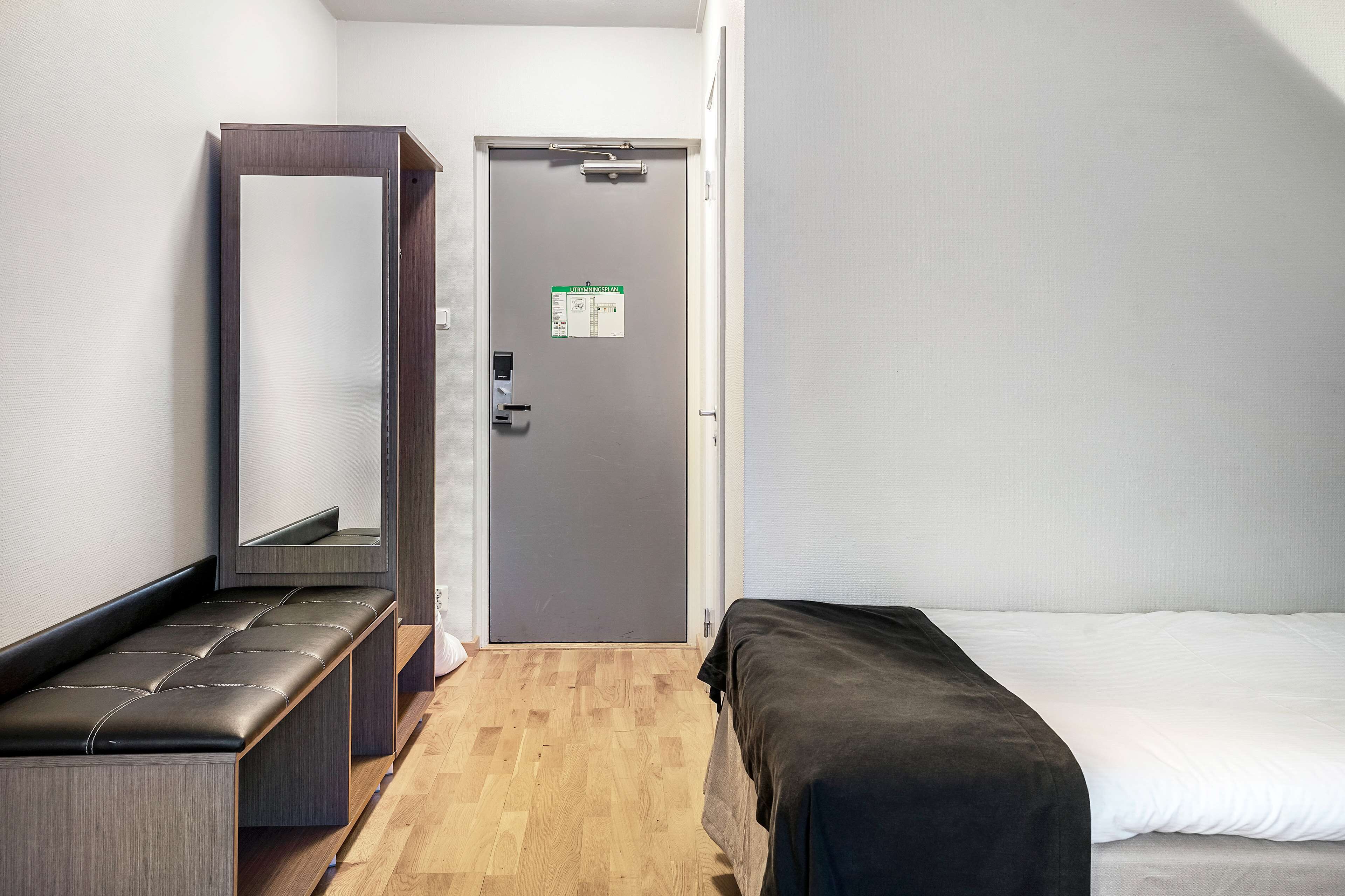 A hotel room with a wooden bench, a bed with a black cover, a door with a sign, and a mirror on the wall.