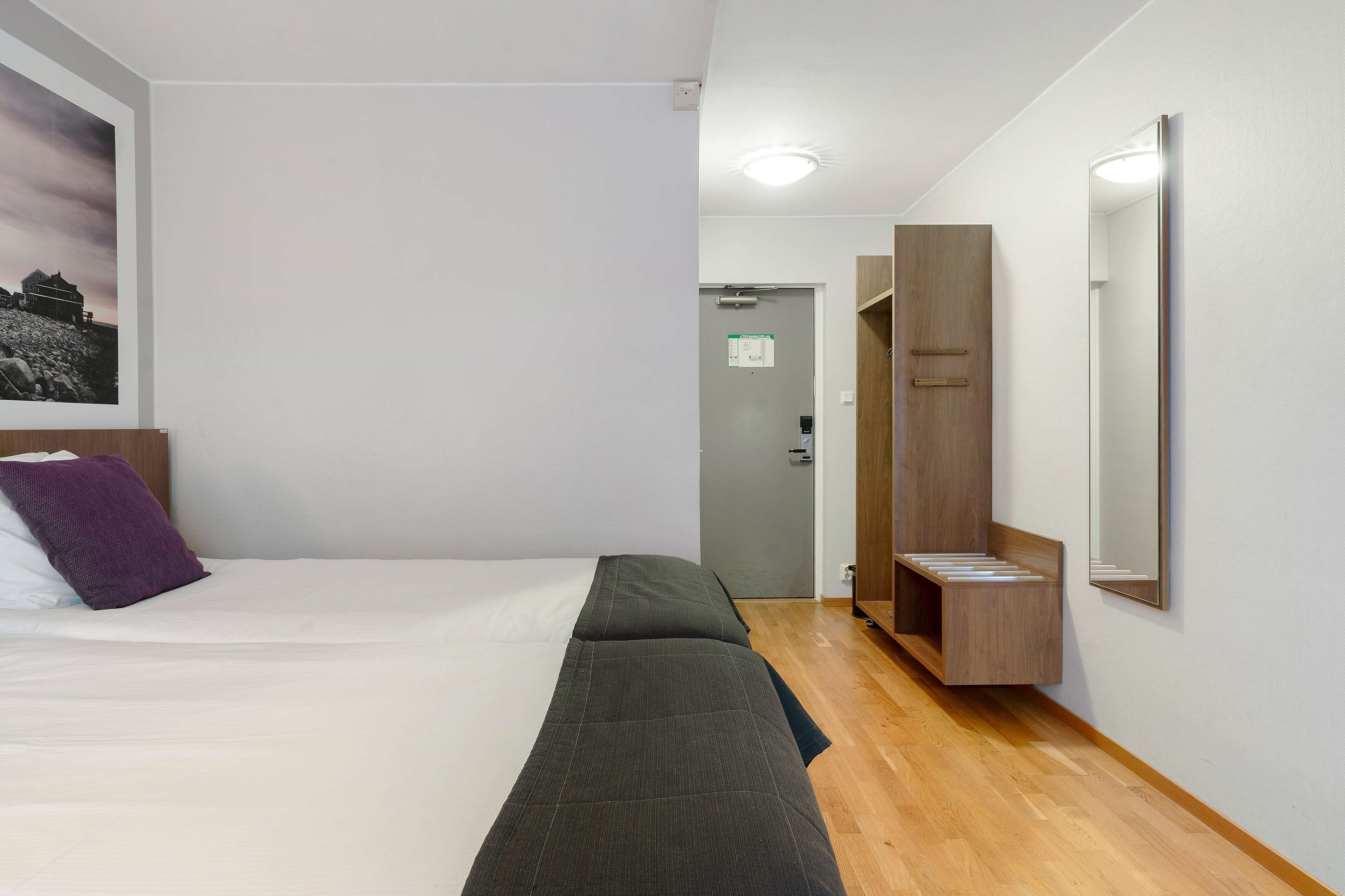 A hotel room with a bed, a wooden bench, a mirror, and a door leading to a bathroom.