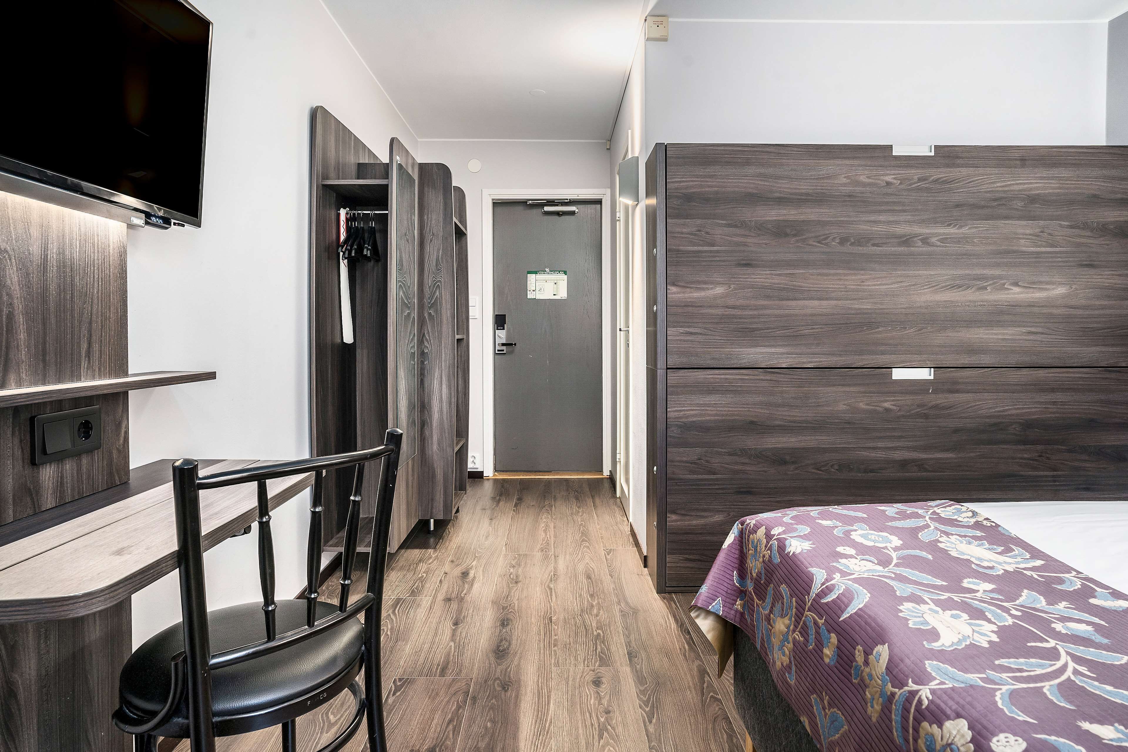 Ett hotellrum med en säng, ett skrivbord, en stol och en tv som är monterad på väggen.
