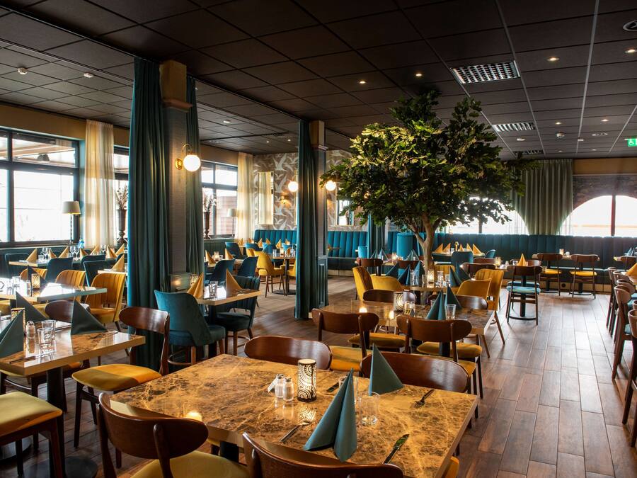 Un restaurante espacioso con numerosas mesas y sillas, grandes ventanales y un árbol central.
