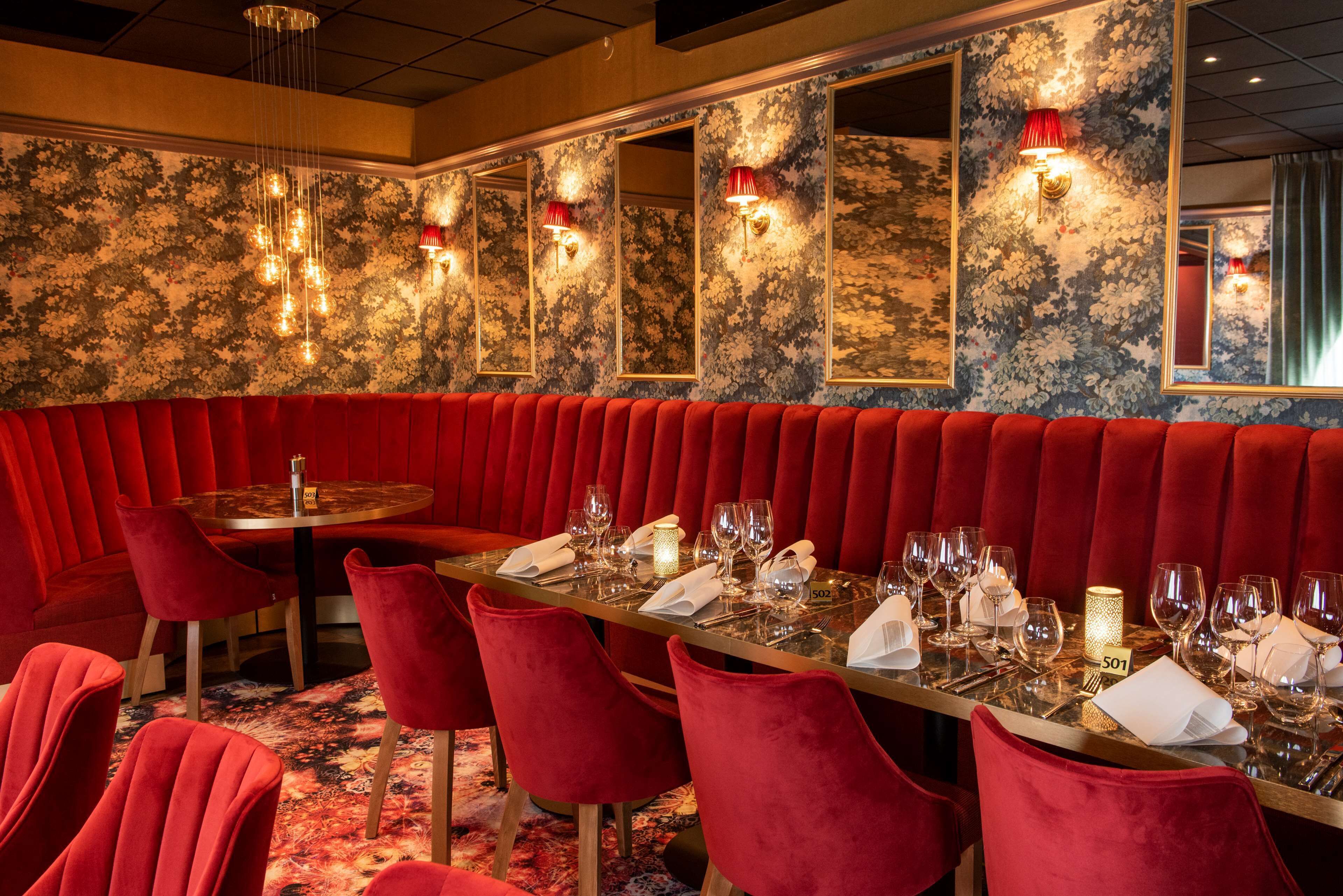 A restaurant with a long red booth seating area, a round table with a number 501, and floral wallpaper.