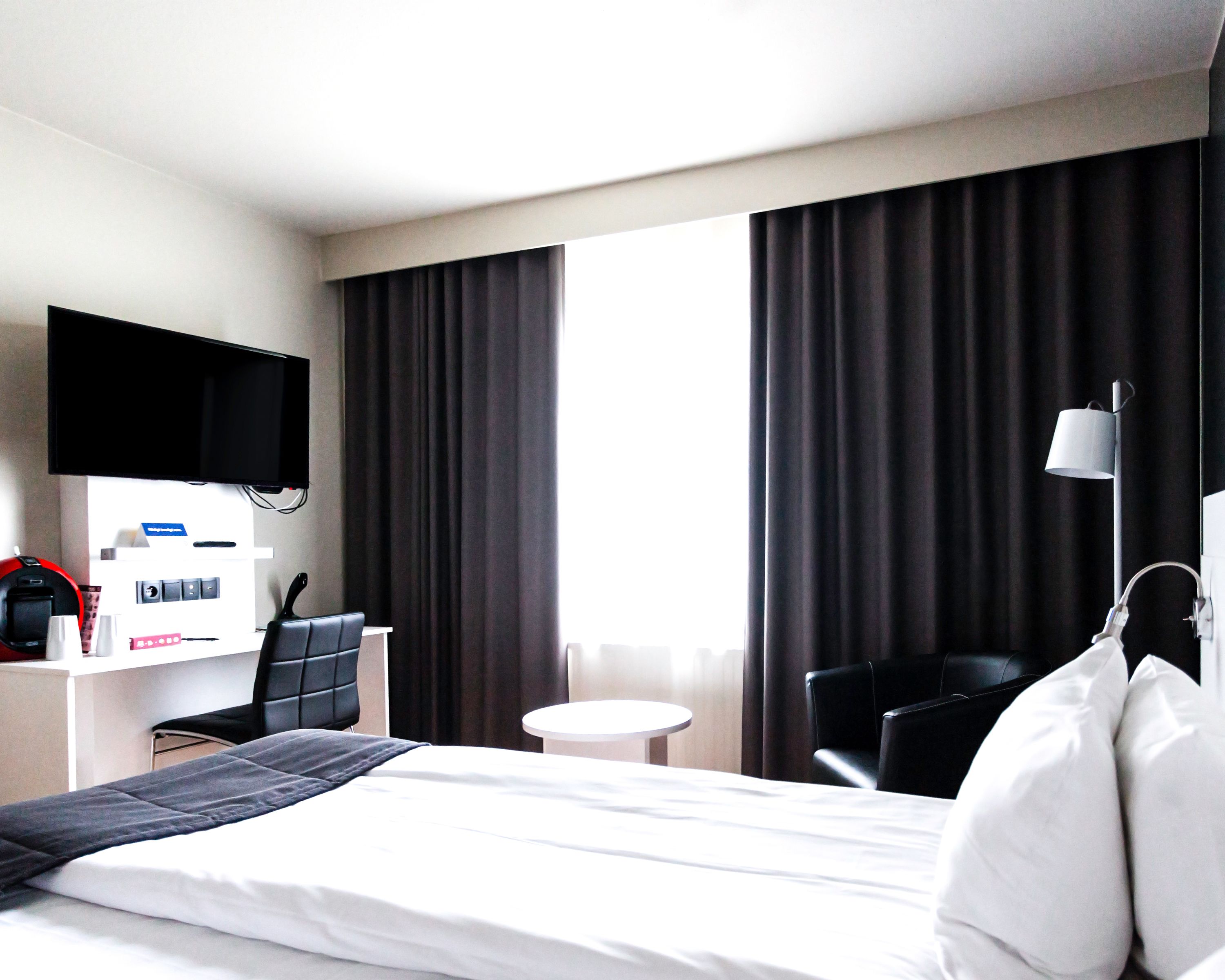 A hotel room with a bed, a desk with a computer, a chair, a TV mounted on the wall, and a window with curtains.