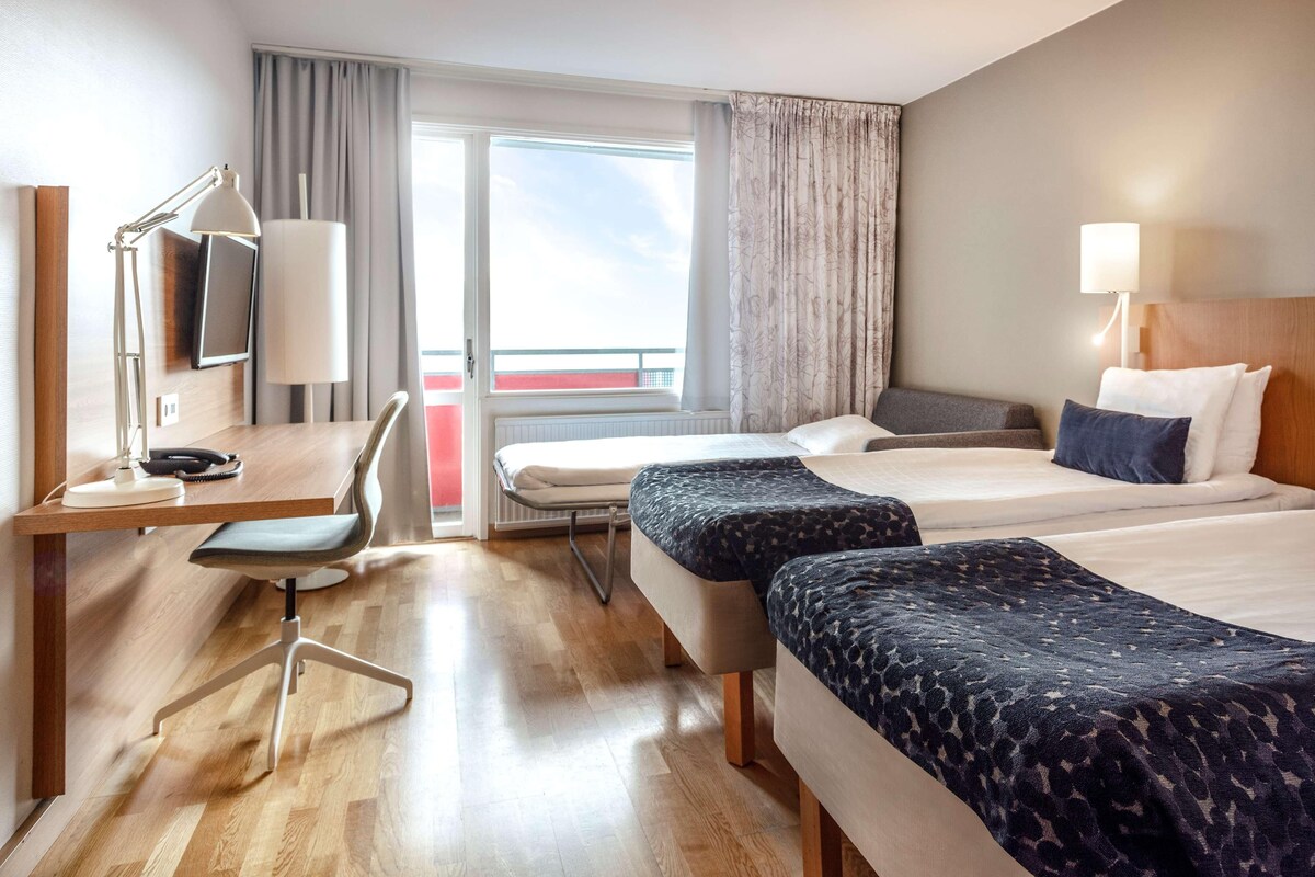 A hotel room with a desk, two beds, a chair, and a window with curtains.