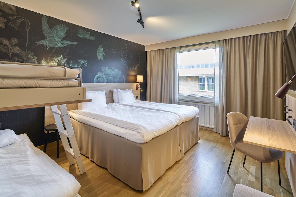 A hotel room with a large bed, a desk, a chair, and a view of a building through the window.