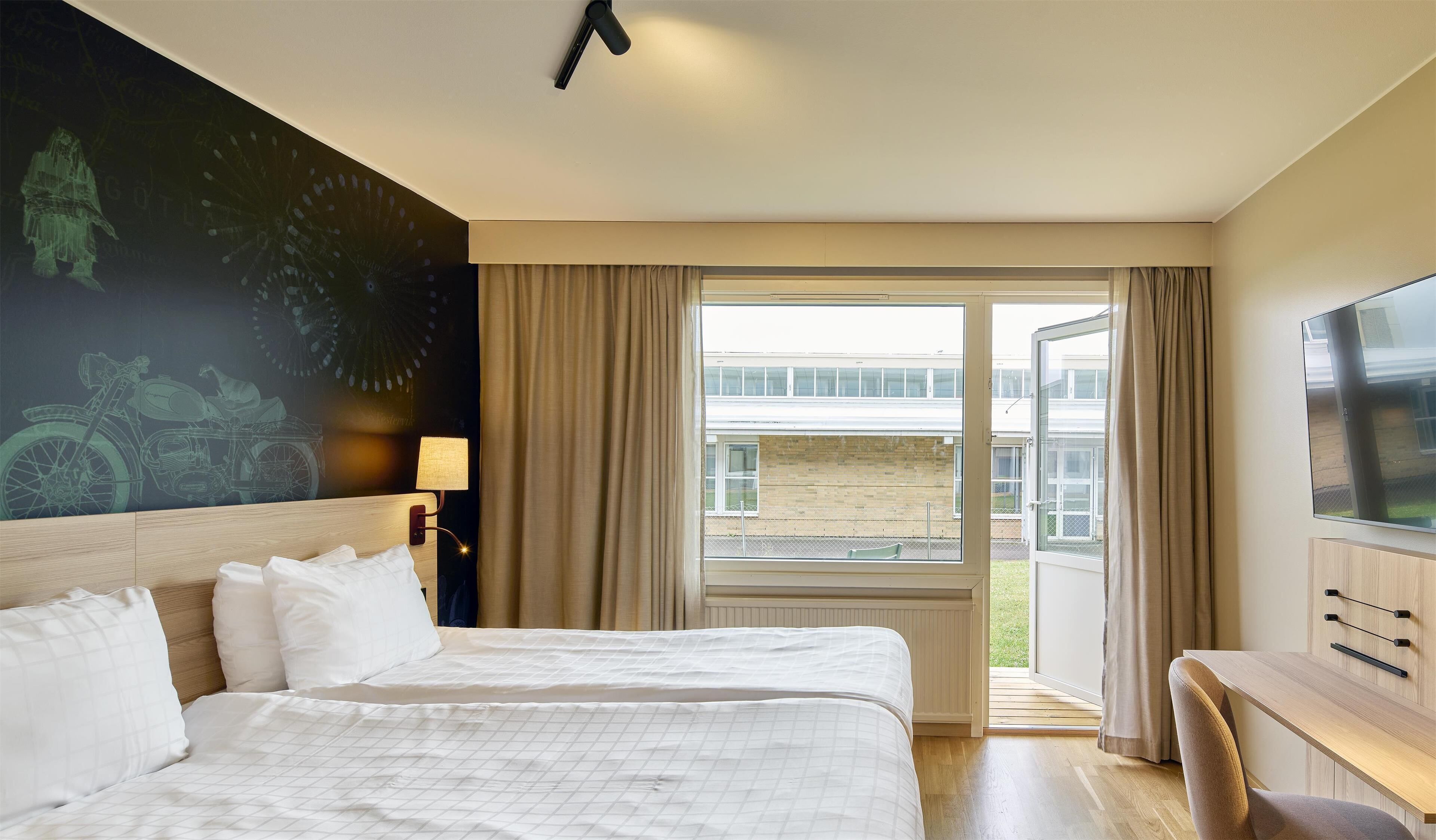 Habitación de hotel con dos camas, televisión, escritorio, lámpara y un ventanal con cortinas.