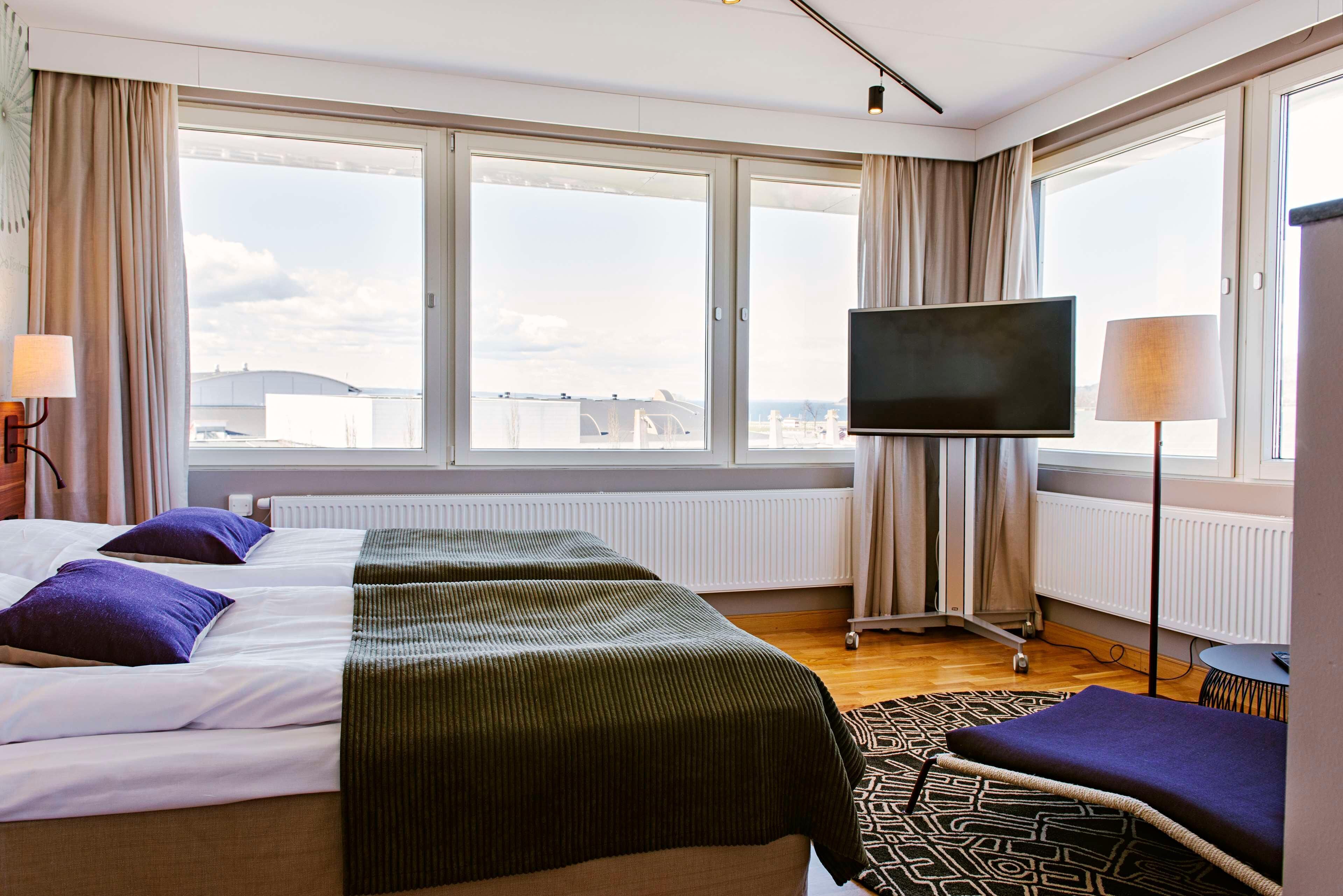 Habitación de hotel con cama, televisión, ventana con cortinas y vista a la ciudad.