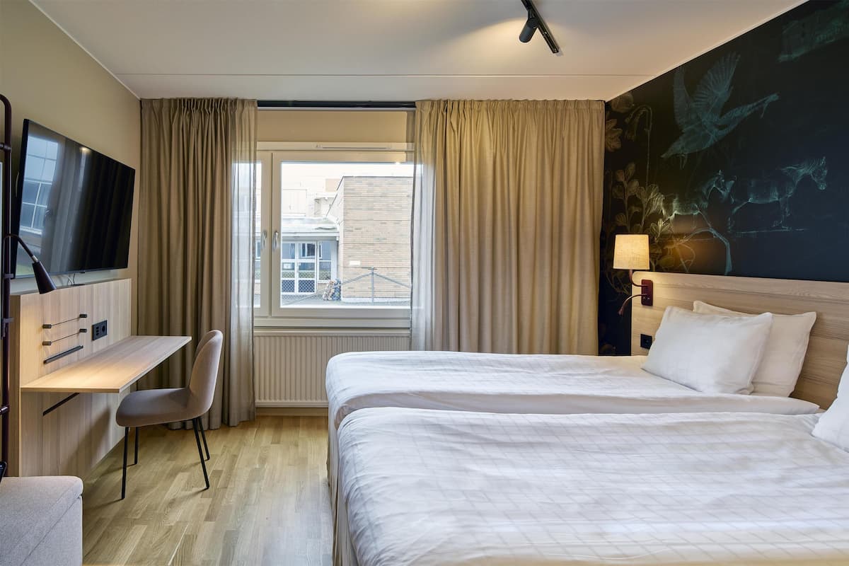 A hotel room with two beds, a desk, a chair, a TV, and a window with curtains.
